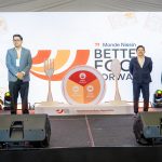 Three presenters stand on a stage in front of a large screen with 'Better Food Forward' branding and playful utensil illustrations, flags on the left.