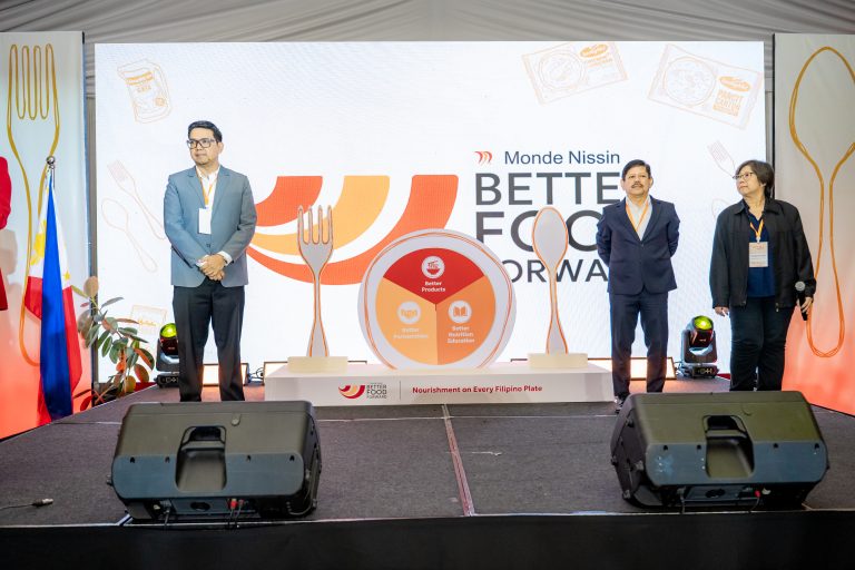 Three presenters stand on a stage in front of a large screen with 'Better Food Forward' branding and playful utensil illustrations, flags on the left.