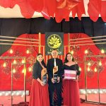 Three award recipients pose on a red-decorated stage, holding trophies and a certificate, smiling at the camera.