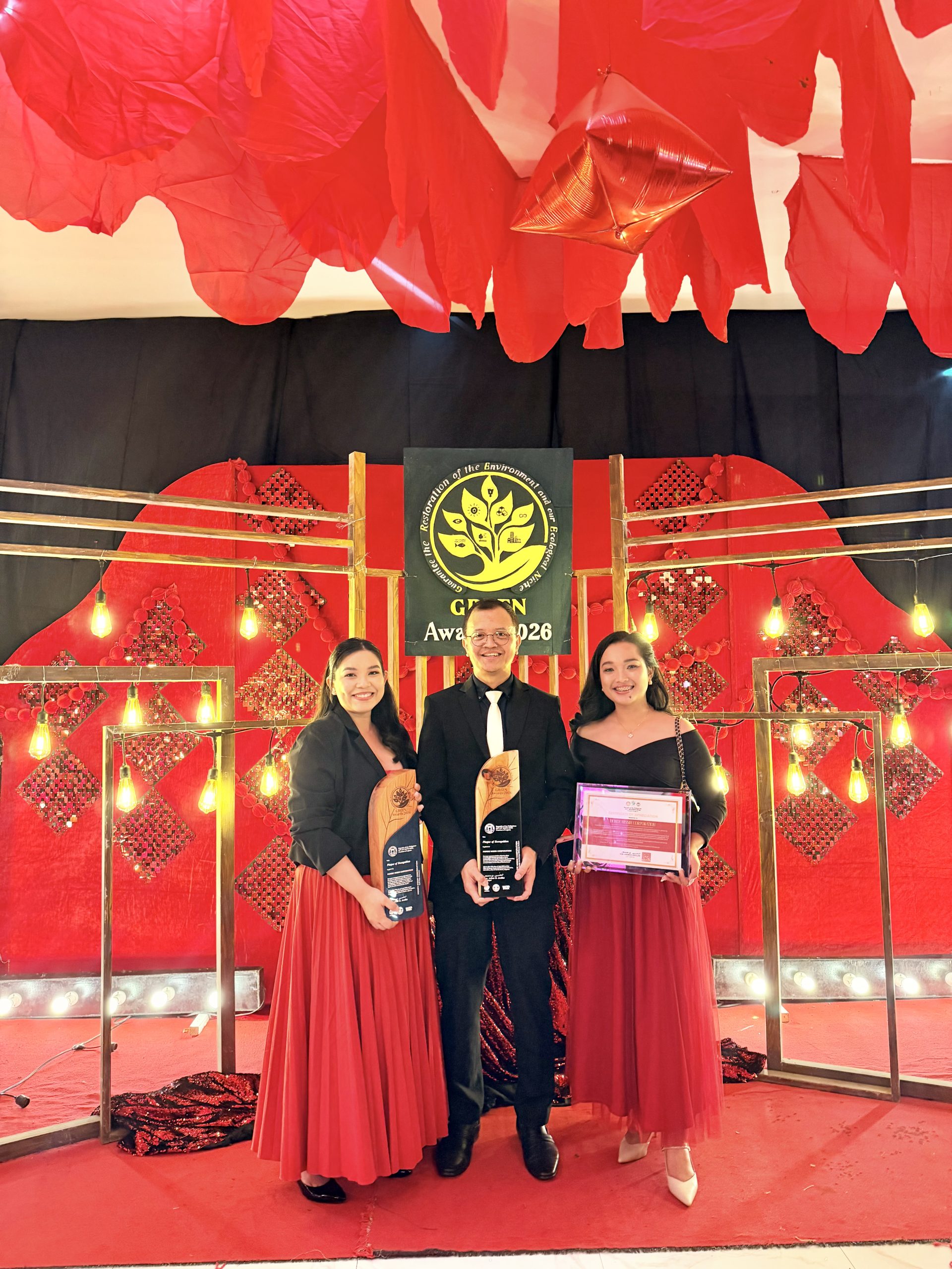 Three award recipients pose on a red-decorated stage, holding trophies and a certificate, smiling at the camera.